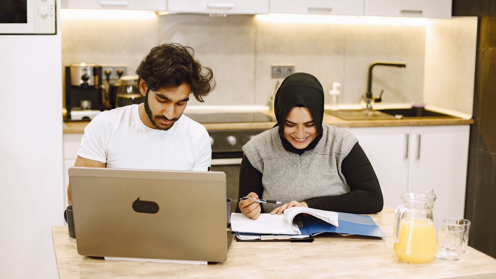 A couple sits at a kitchen table with a laptop and an open folder of paperwork.