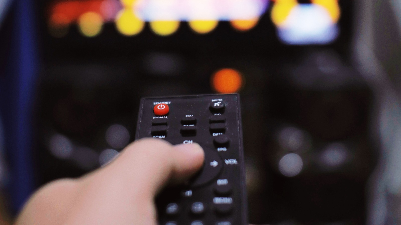 A hand points a television remote toward a softly blurred screen in a dim living room.