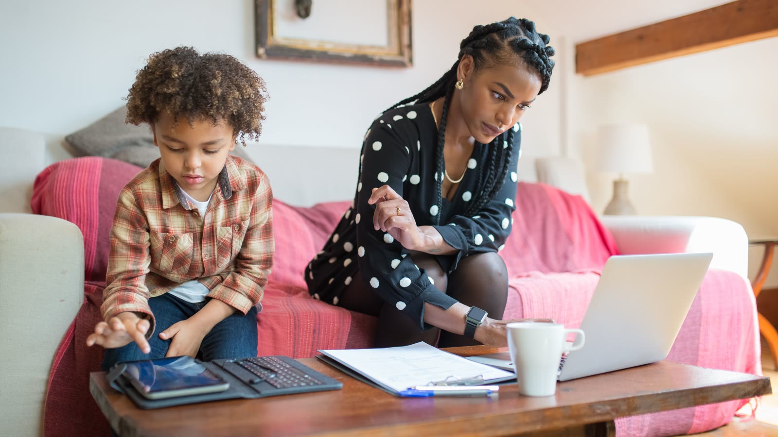 A woman works on a laptop at a living-room table while a child uses a tablet beside her.