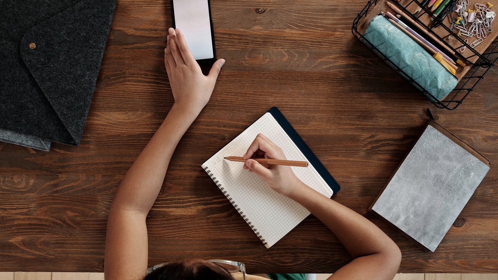 A child writes in a notebook beside a smartphone on a wooden desk.