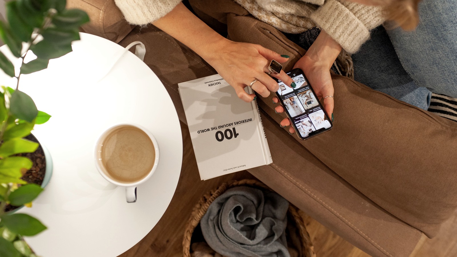 A person uses a smartphone beside a book and coffee in a softly lit living room.