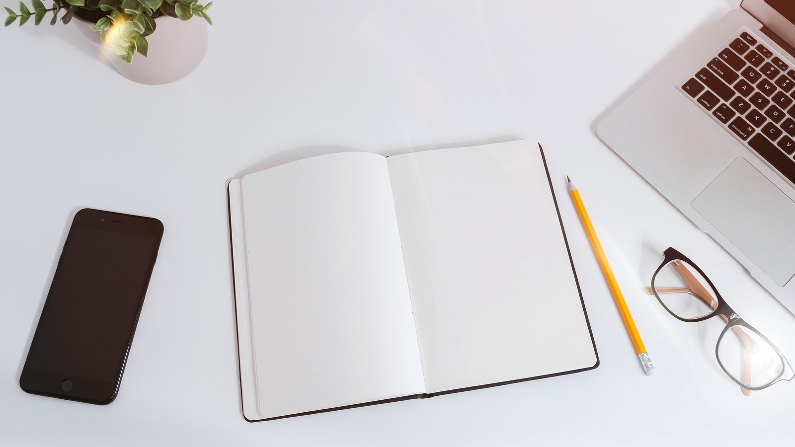 An open blank notebook sits between a smartphone and a laptop on a white desk.